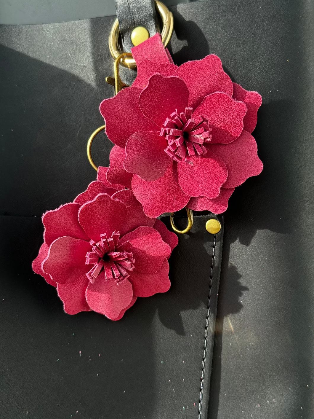 A small leather flower accessory in old rose color, attached to a black strap with a lobster clasp and a split ring, showing the texture and color of the leather.