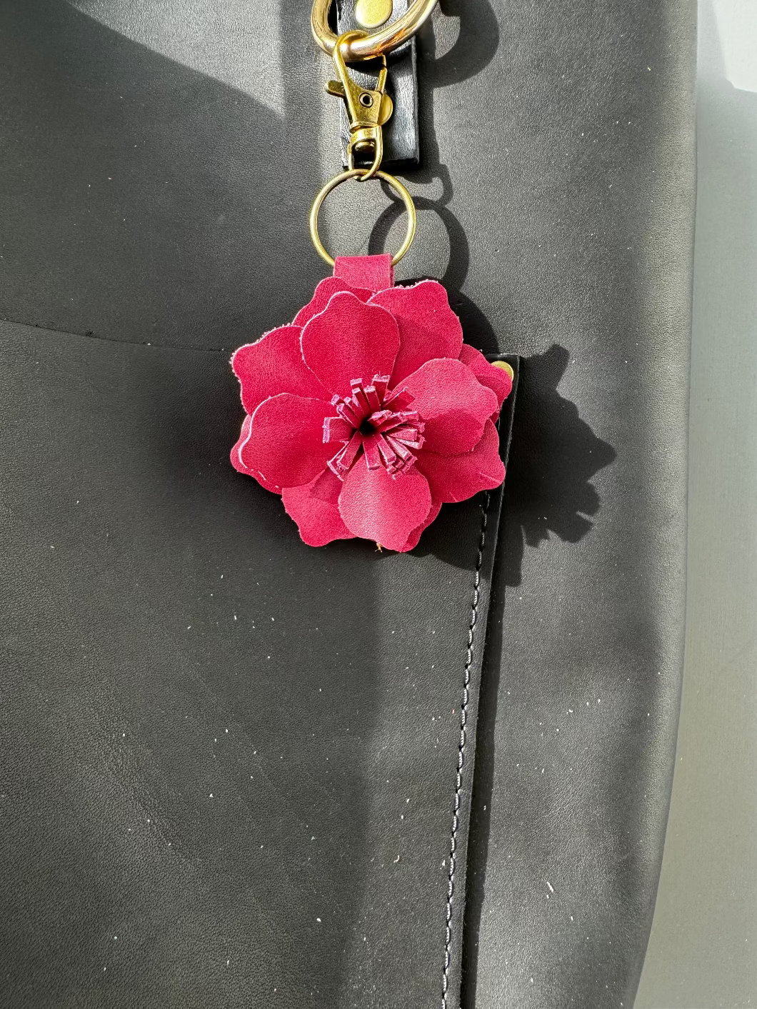 A small leather flower accessory in old rose color, attached to a black strap with a lobster clasp and a split ring, showing the texture and color of the leather.