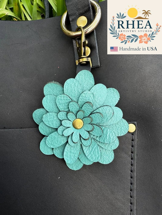 Teal flower-shaped leather purse charm attached to a black bag with RHEA Artistry Studios branding.