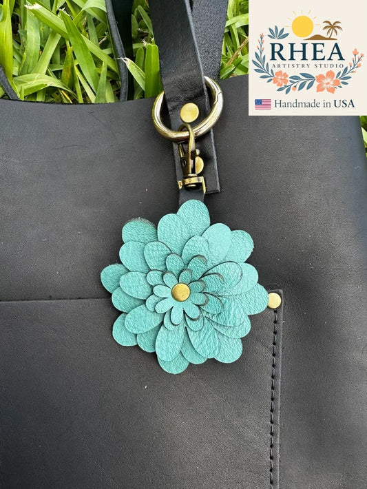 Teal flower-shaped leather bag charm attached to a black bag with RHEA Artistry Studio branding.