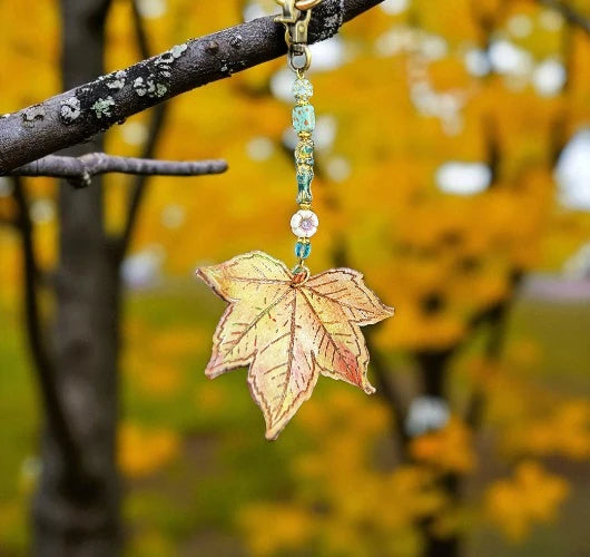 Leaf-shaped keychain with beads hanging from a branch against a blurred autumn background