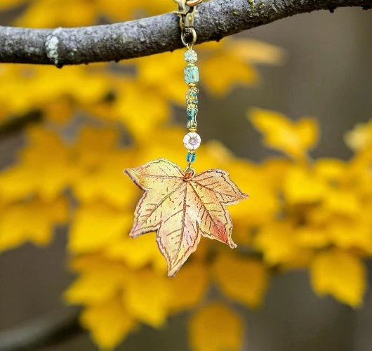 Leaf-shaped keychain with beads hanging from a branch with yellow leaves in the background