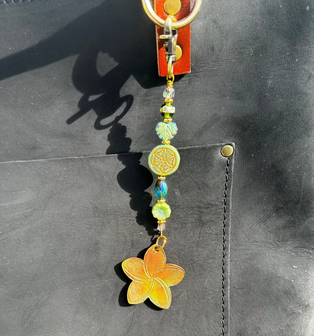 A handcrafted plumeria charm made with leather and solid brass hardware, featuring czech glass accents, attached to a keychain.