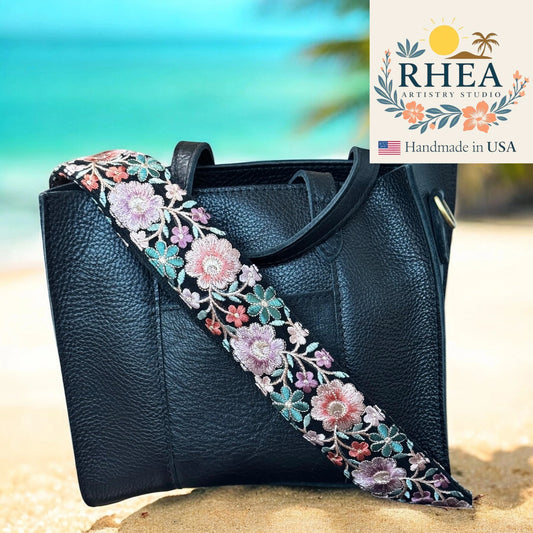 Black handbag with a floral purse strap on a beach background, featuring RHEA Artistry Studio logo.