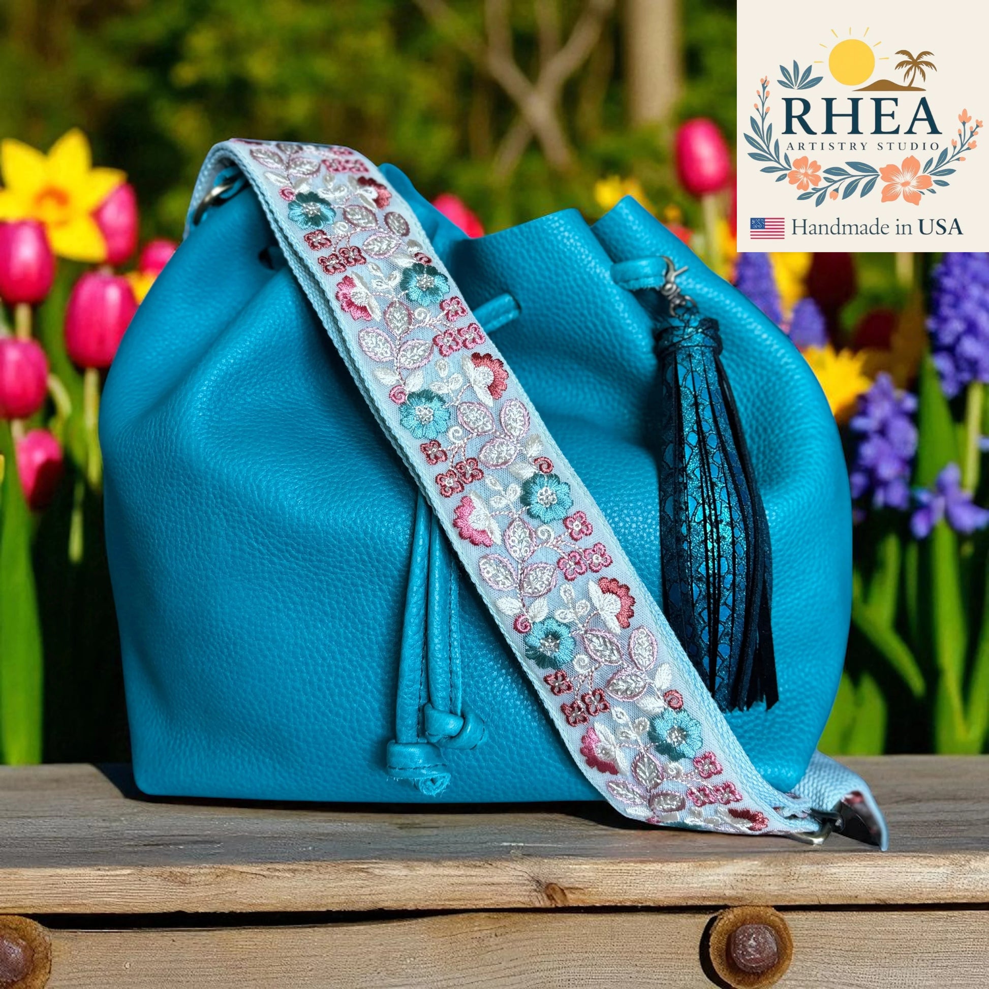 Blue handbag with a decorative strap in front of flowers, featuring the RHEEA brand.