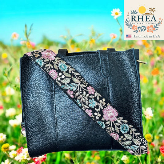 Black handbag with floral strap against a colorful flower field background, featuring the RHEA Artistry Studio logo.