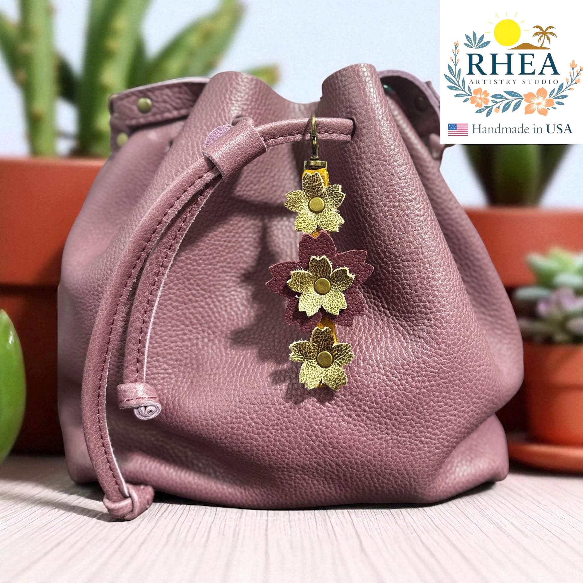 Pink leather bag with floral charm on a blurred background, featuring the RHEEA brand.