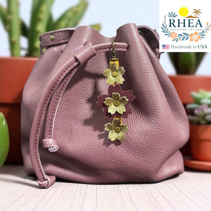 Pink leather bag with floral charm on a blurred background, featuring the RHEEA brand.