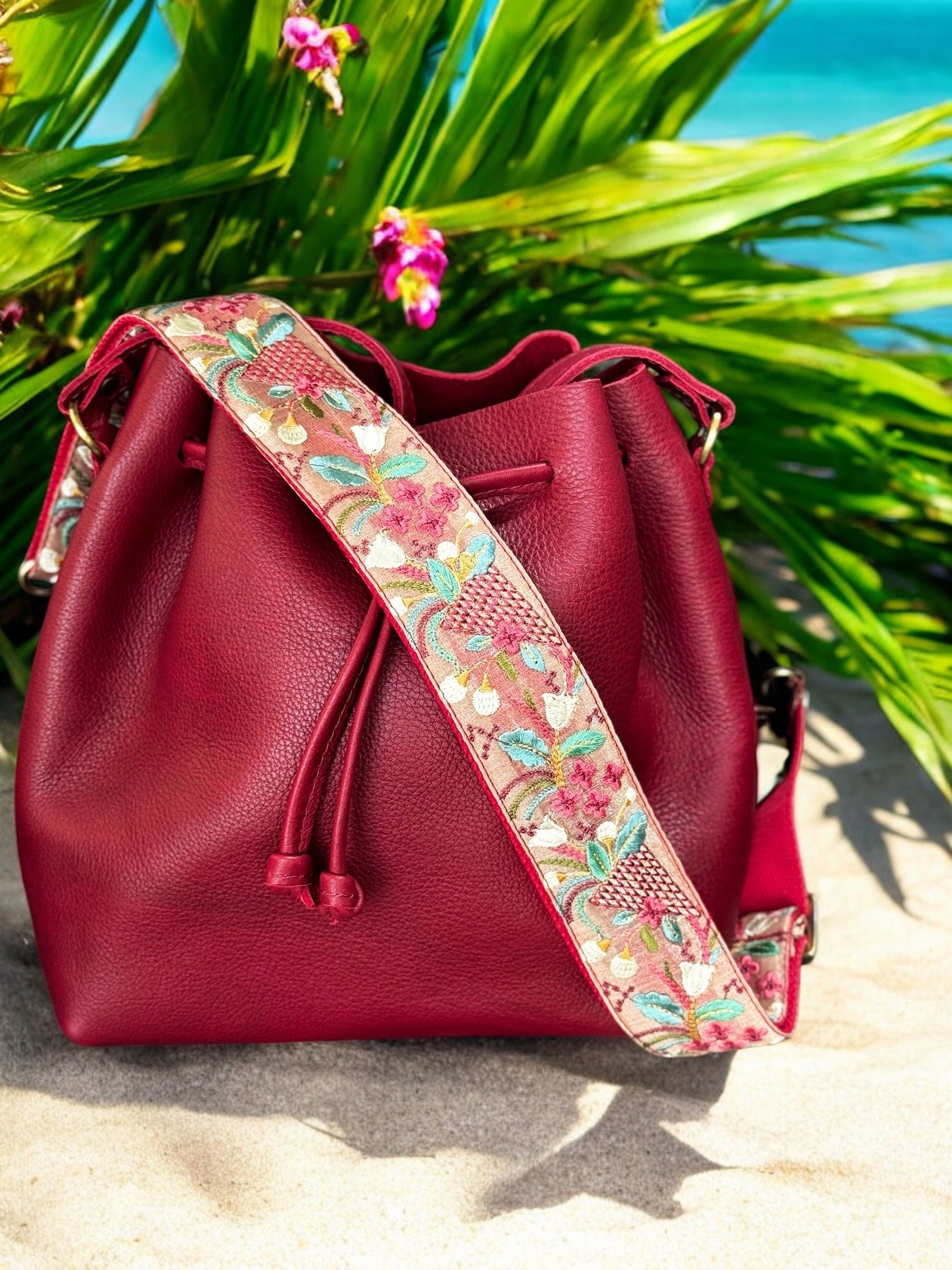 Embroidered strap with autumnal florals and wheat-inspired motifs in ivory, teal, and gold on a coral-pink background on a sandy beach with palm leaves
