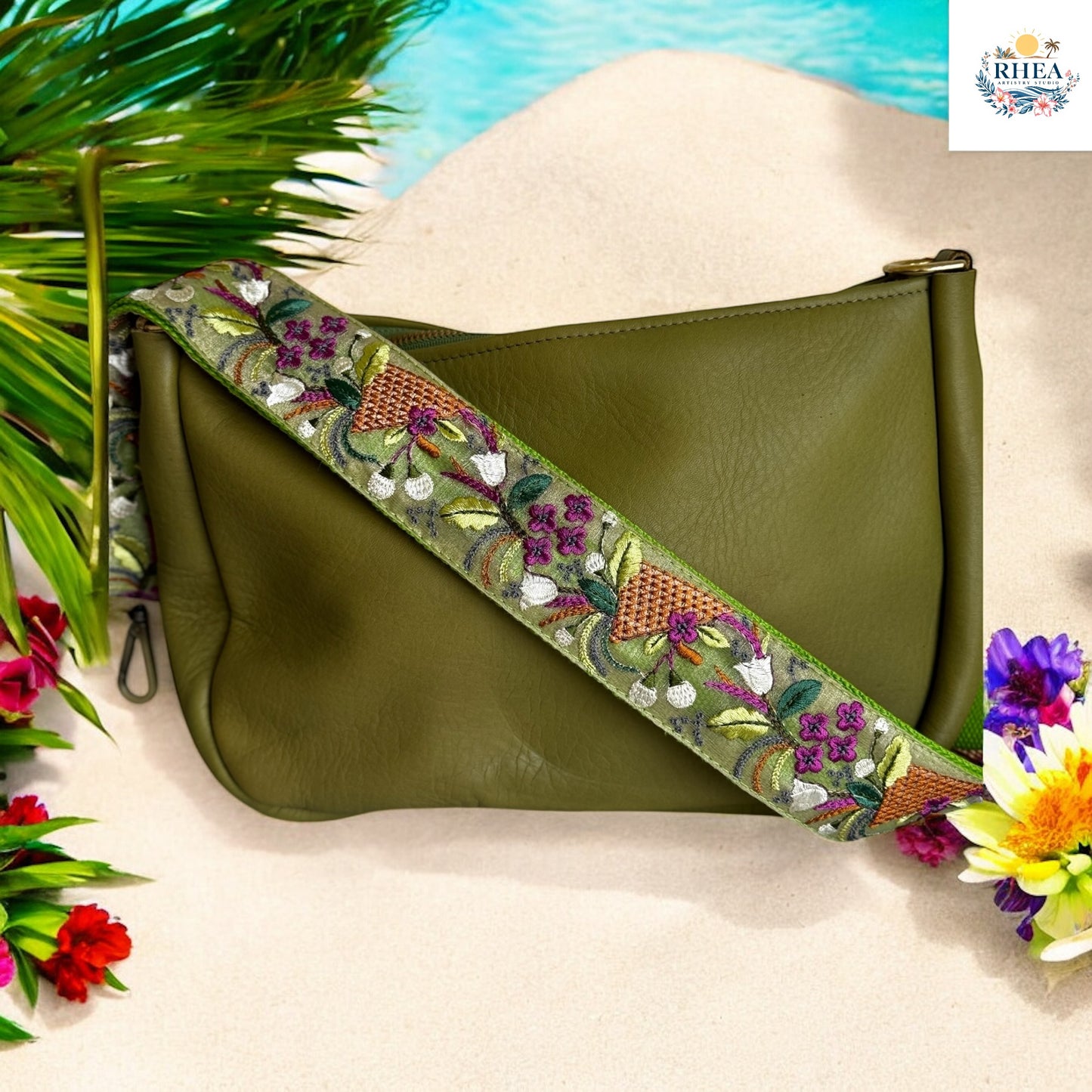 Embroidered strap with deep green base featuring plum and white flowers, copper basket weave embroidery, and leafy vine details, displayed on an olive leather bag on a sandy beach