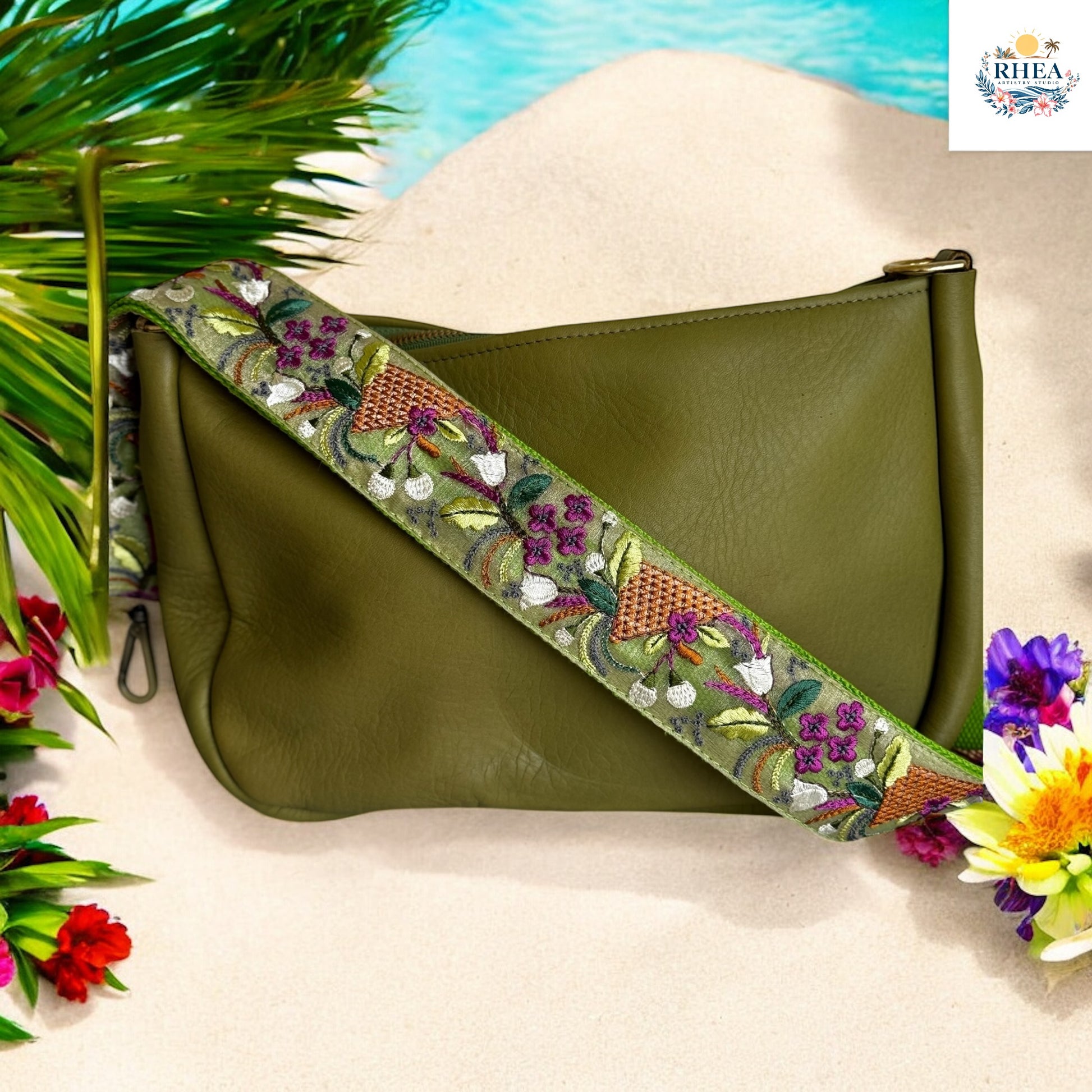 Embroidered strap with deep green base featuring plum and white flowers, copper basket weave embroidery, and leafy vine details, displayed on an olive leather bag on a sandy beach