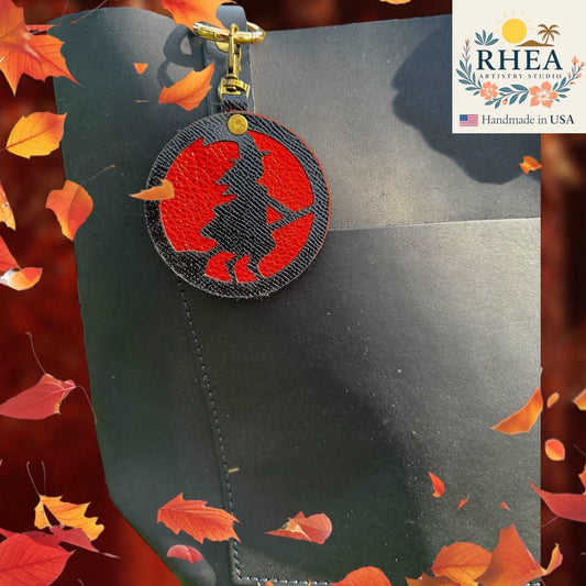 Black leather product with a red and black embroidered witch leather purse charm on a background with autumn leaves and RHEA Artisan Studio branding.