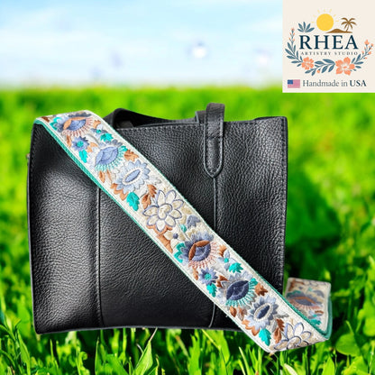 Black leather bag with an embroidered purse strap on a grassy background, featuring the RHEA Artistry Studios logo.