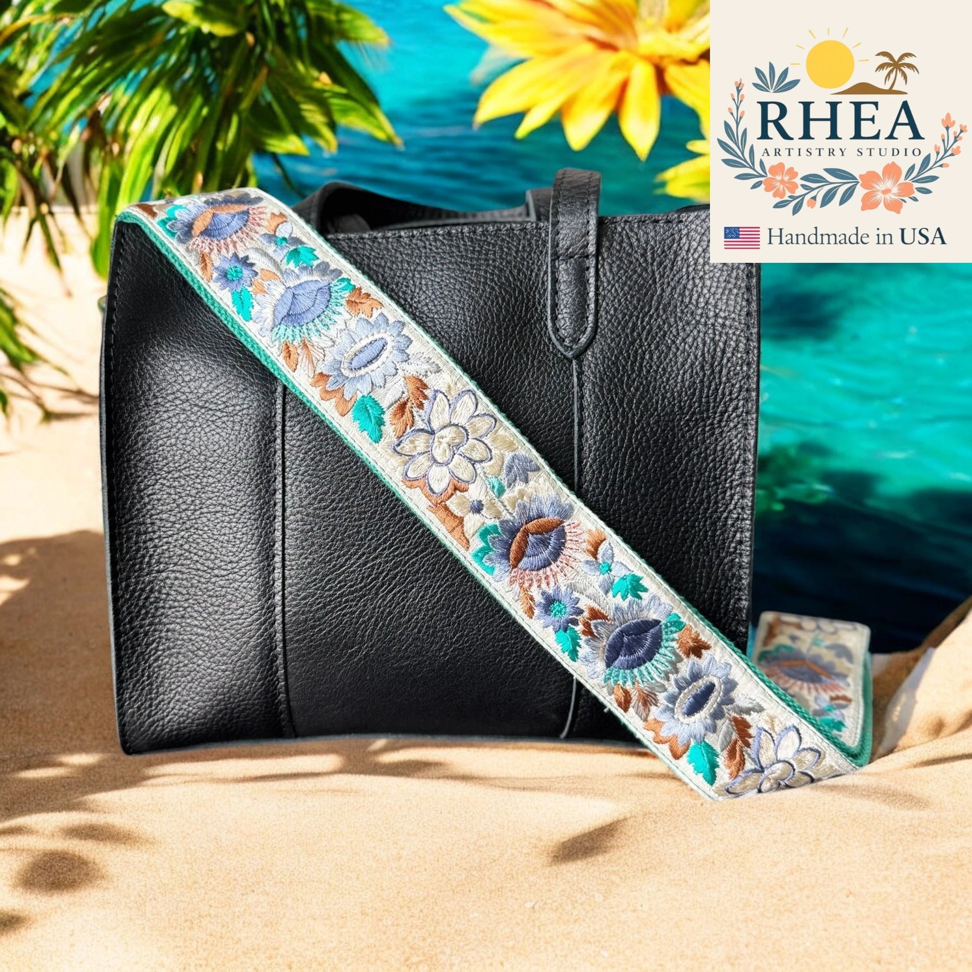 Black handbag with a floral purse strap on sand, flowers, and RHEA Artistry Studio logo.