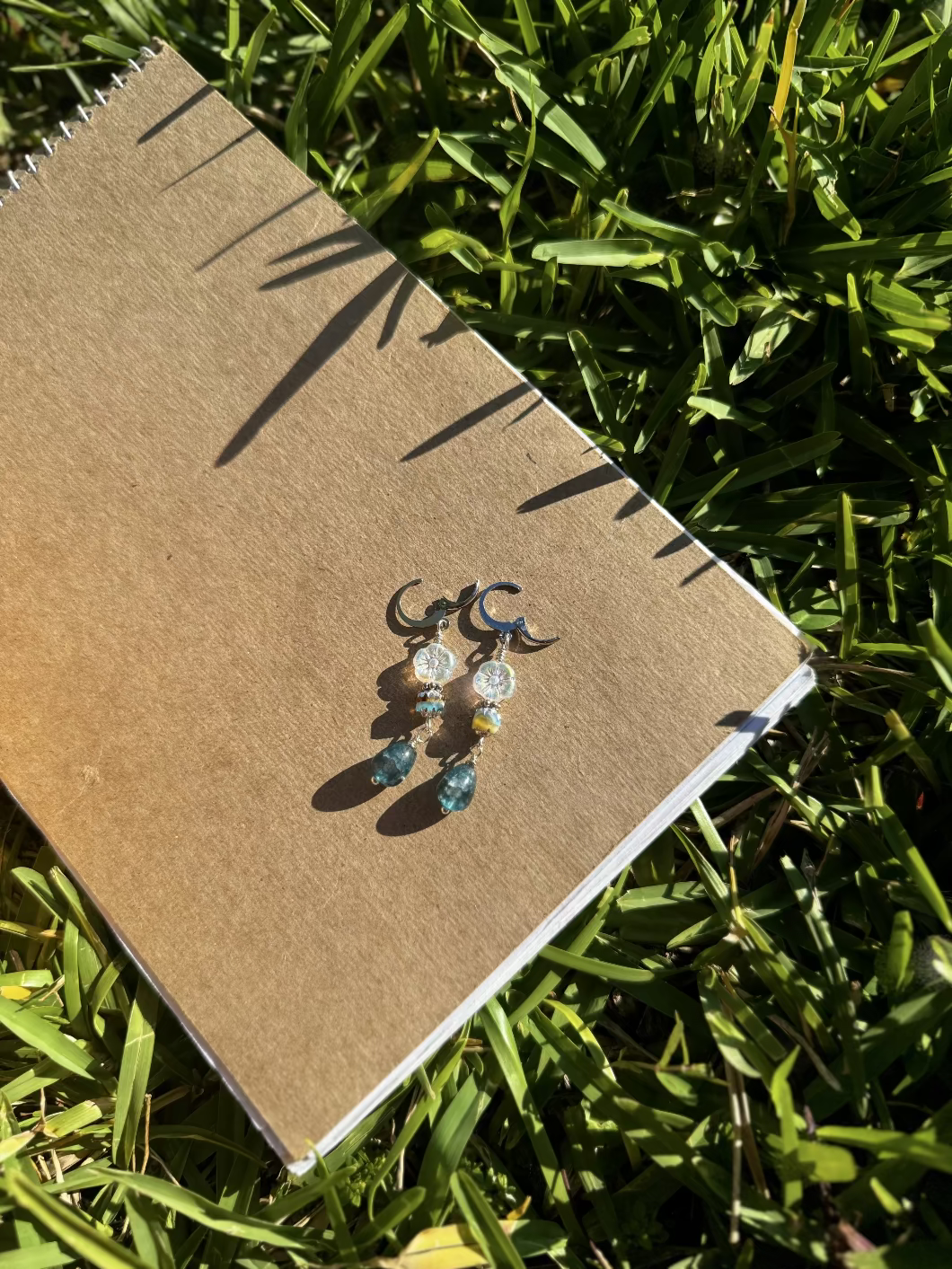 A pair of silver drop earrings with blue and clear embellishments, displayed on a brown notebook outdoors.