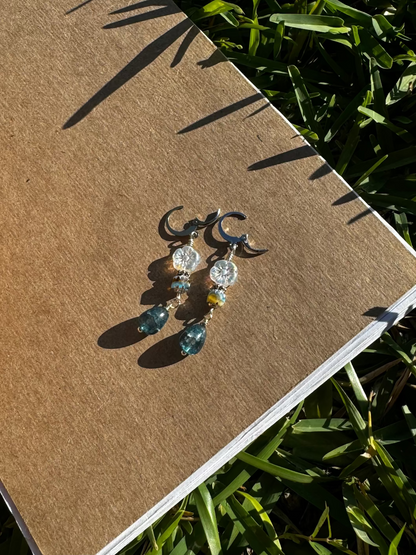 A pair of silver drop earrings with blue and clear embellishments, displayed on a brown notebook outdoors.