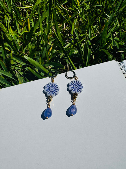 A pair of blue glass drop earrings with gold plated brass findings, displayed on a flat surface with grass and a clear sky in the background.