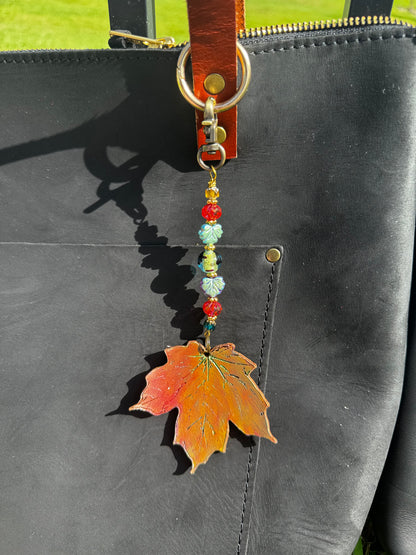 A handcrafted maple leaf charm made of vegetable tan leather with a brass hardware and multicolored beads, designed to be worn on a bag.