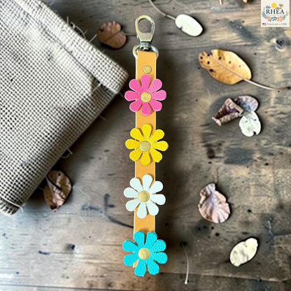 Rhea Artistry Studio handmade leather daisy wristlet keychain with pink, yellow, and blue flowers attached to a tan leather strap with brass rivets.