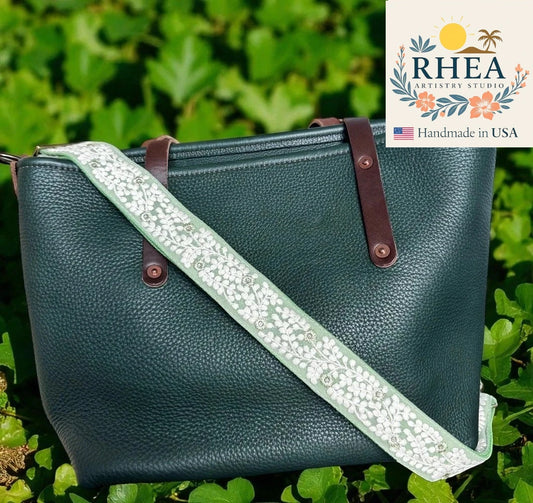 Green leather handbag with a floral strap on a green leafy background, featuring the RHEA Artistry Studio logo.