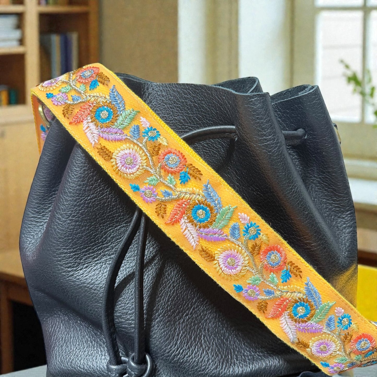  Adjustable length detail showing slider on embroidered strap, Black leather bag with a colorful floral strap in an indoor setting