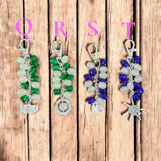 Four beaded keychains with silver charms on a wooden background, labeled Q, R, S, T.