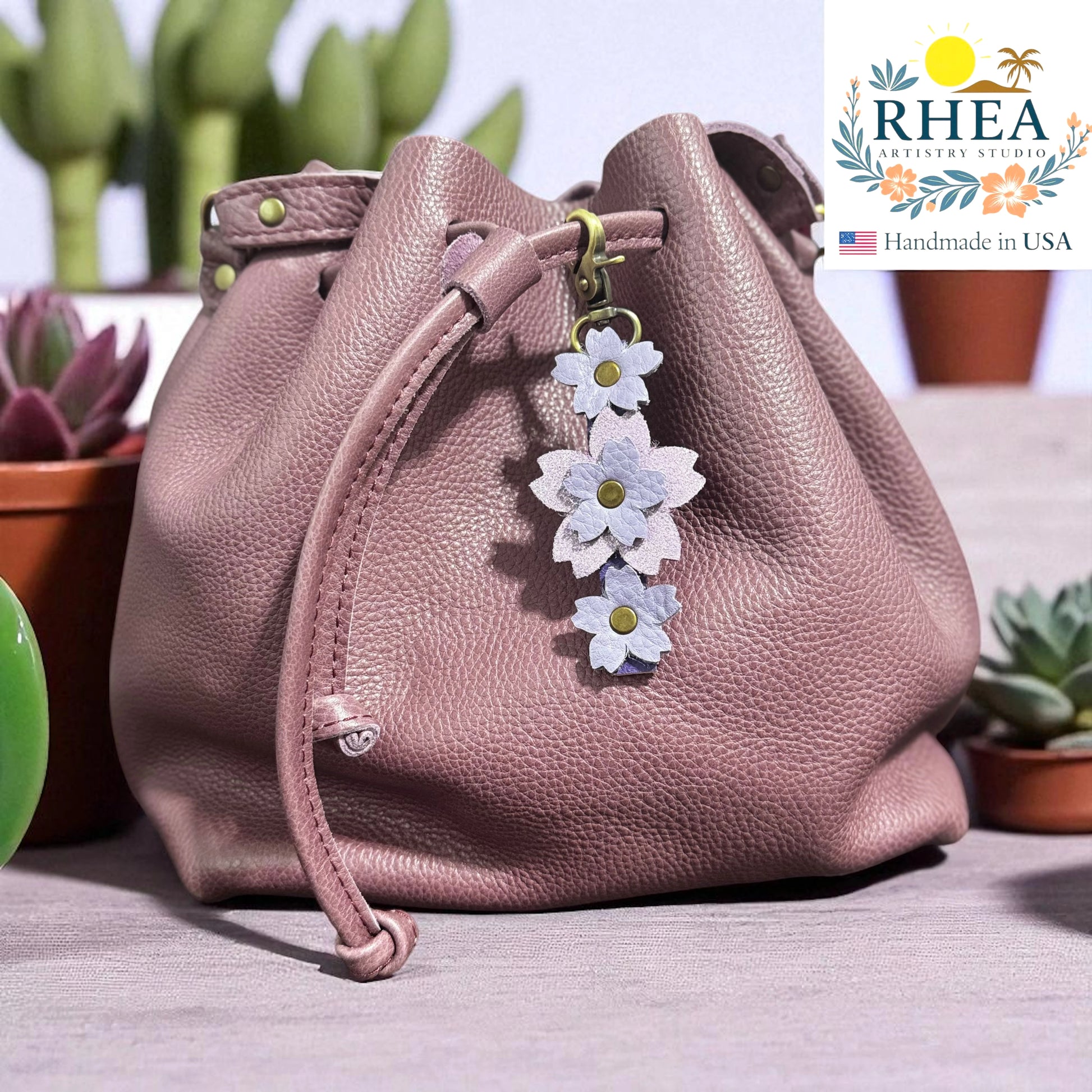 Lavender Sparkles leather bag with floral charm on a surface with plants in the background, featuring the RHEA brand.