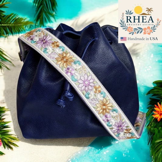 Navy blue handbag with floral purse strap on a tropical background, featuring the RHEA Artistry Studio logo.
