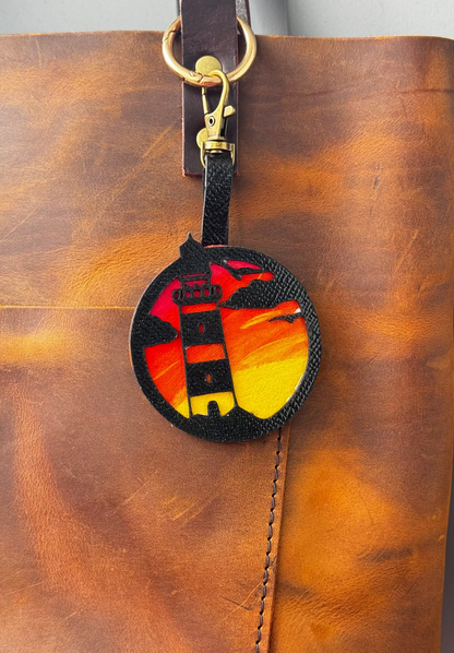 A hand-painted leather charm with a lighthouse design, attached to a brown leather bag with a brass clasp.