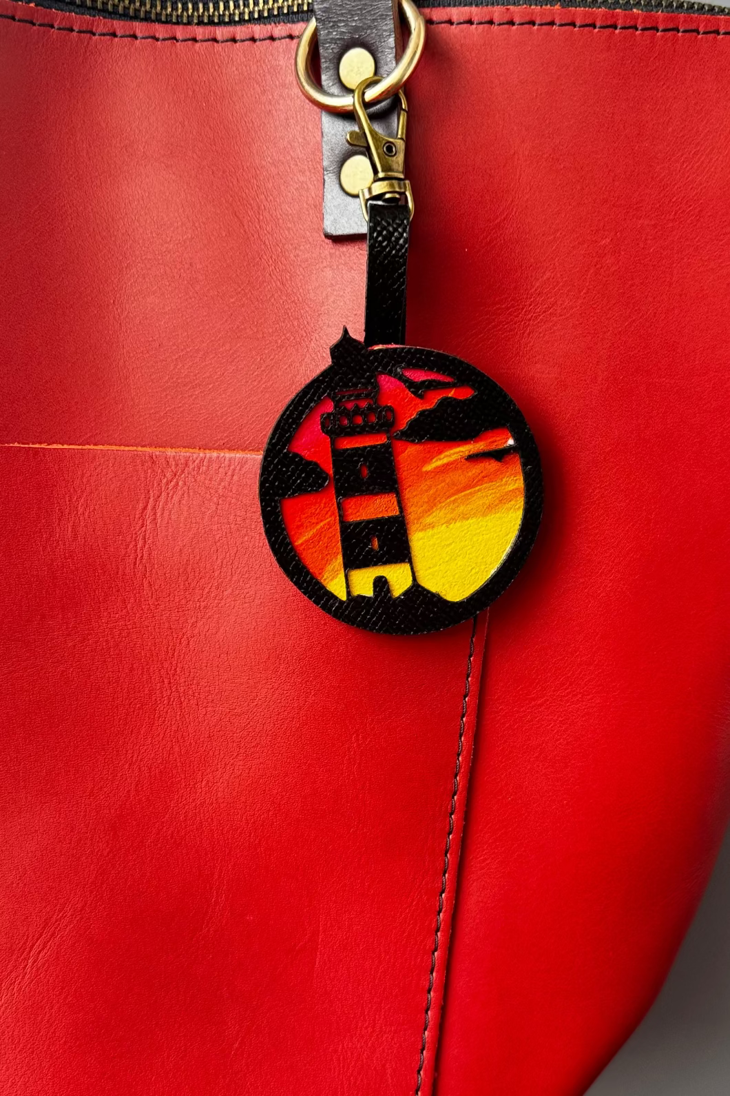 A hand-painted leather charm with a lighthouse design, attached to a red leather bag with a brass clasp.