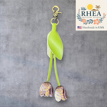 Green bird-shaped leather purse charm with decorative elements on a gray background, featuring the RHEA Artistry Studio logo.