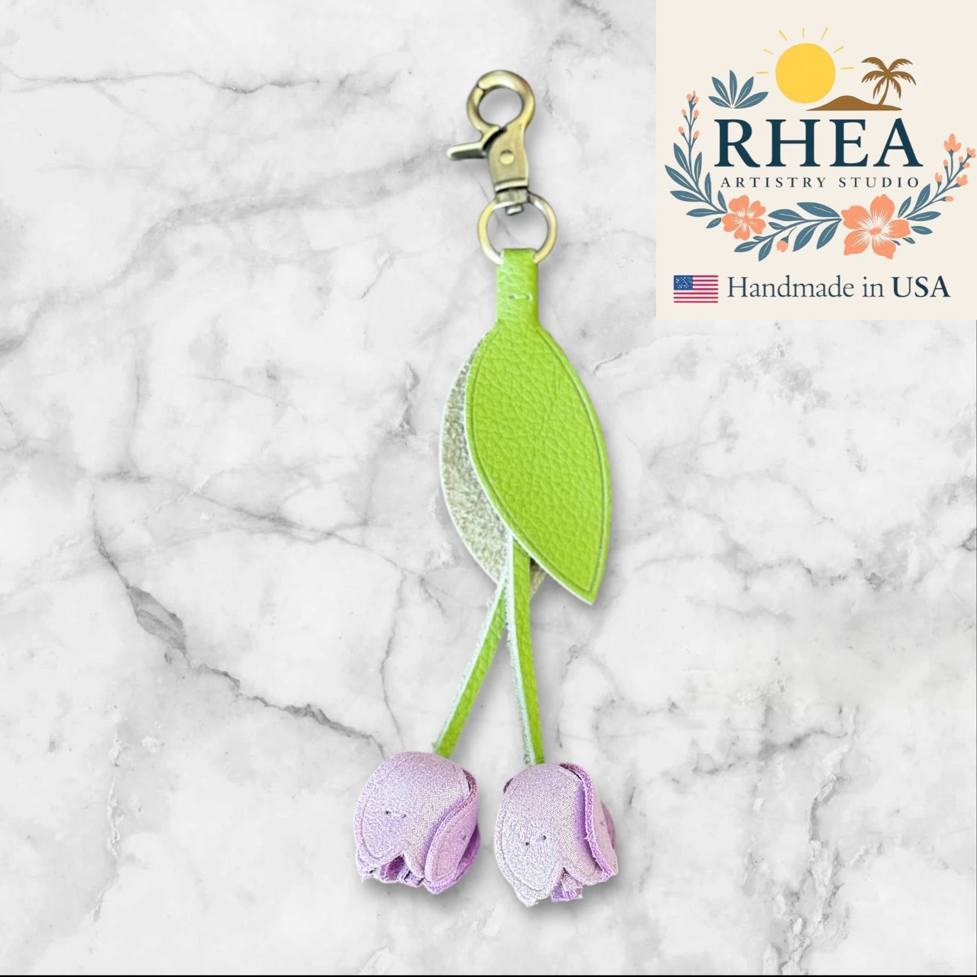 leather purse charm with green leaves and purple flowers on a marble background, featuring RHEA Artistry Studio logo.