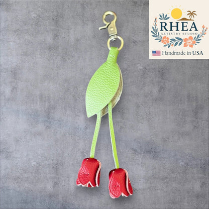 Green leaf-shaped leather purse charm with red flowers on a gray background, branded 'RHEA Artistry Studio'.