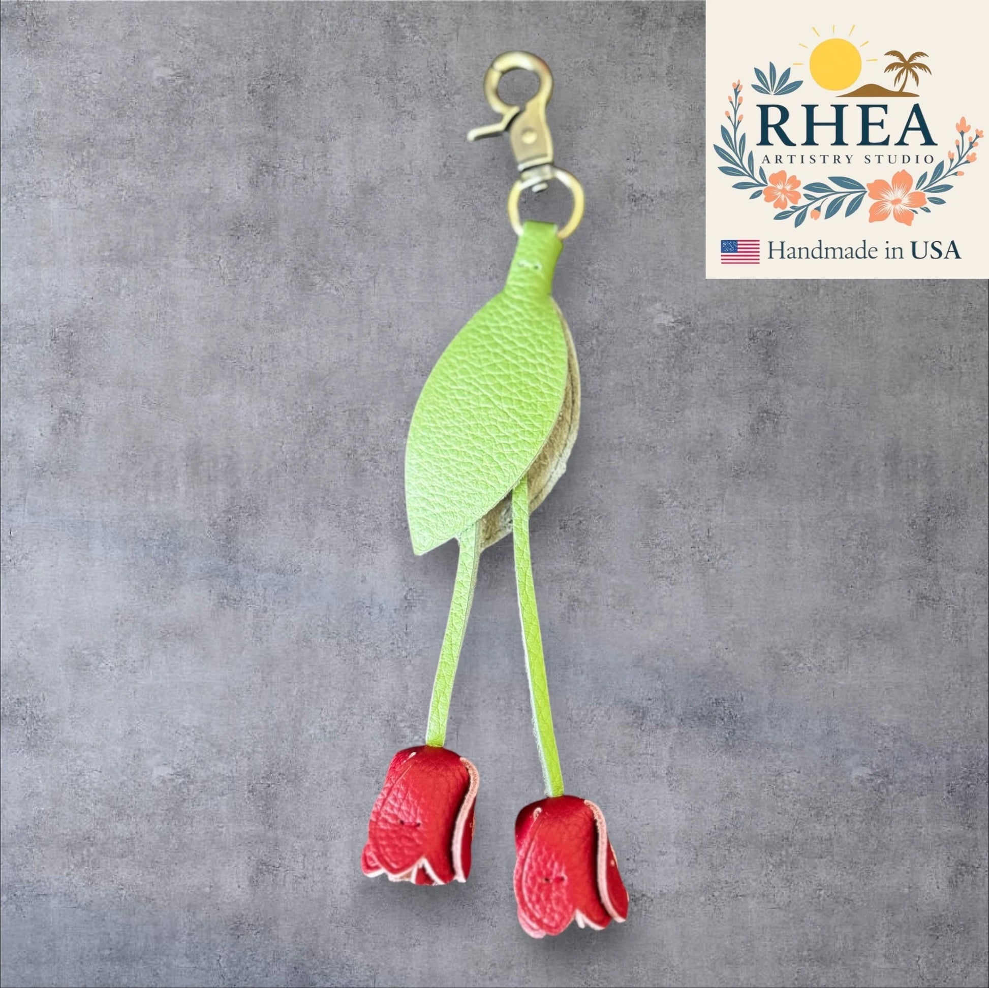 Green leaf-shaped leather purse charm with red flowers on a gray background, branded 'RHEA Artistry Studio'.