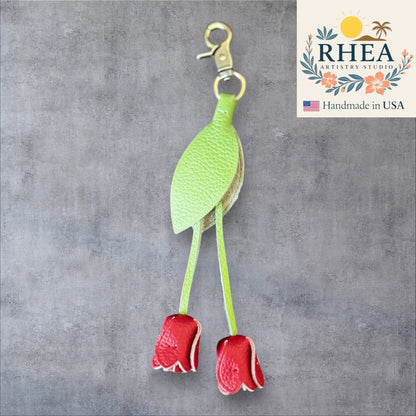 Green leaf-shaped leather purse charm with red flowers on a gray background, branded 'RHEA Artistry Studio'.