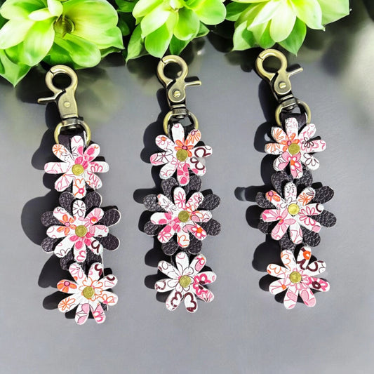A handcrafted leather charm featuring layered daisy motifs in printed and solid leather petals, connected vertically with a gold center and antique brass lobster clasp, styled against a soft neutral backdrop with floral accents.