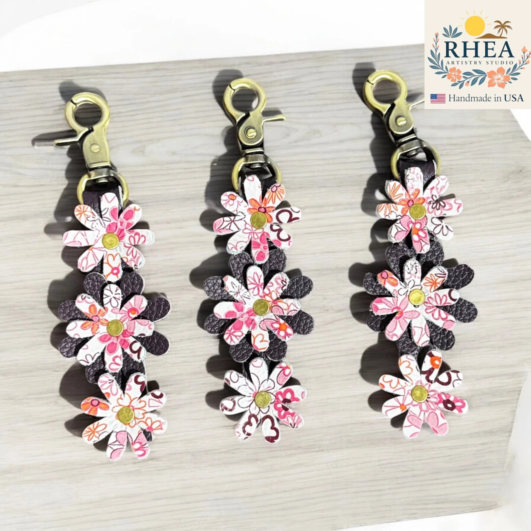 Floral leather bag charms with RHEA Artistry Studio branding on a light wooden surface.