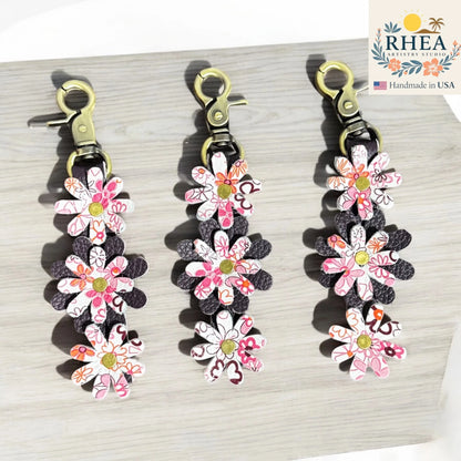 Floral leather bag charms with RHEA Artistry Studio branding on a light wooden surface.