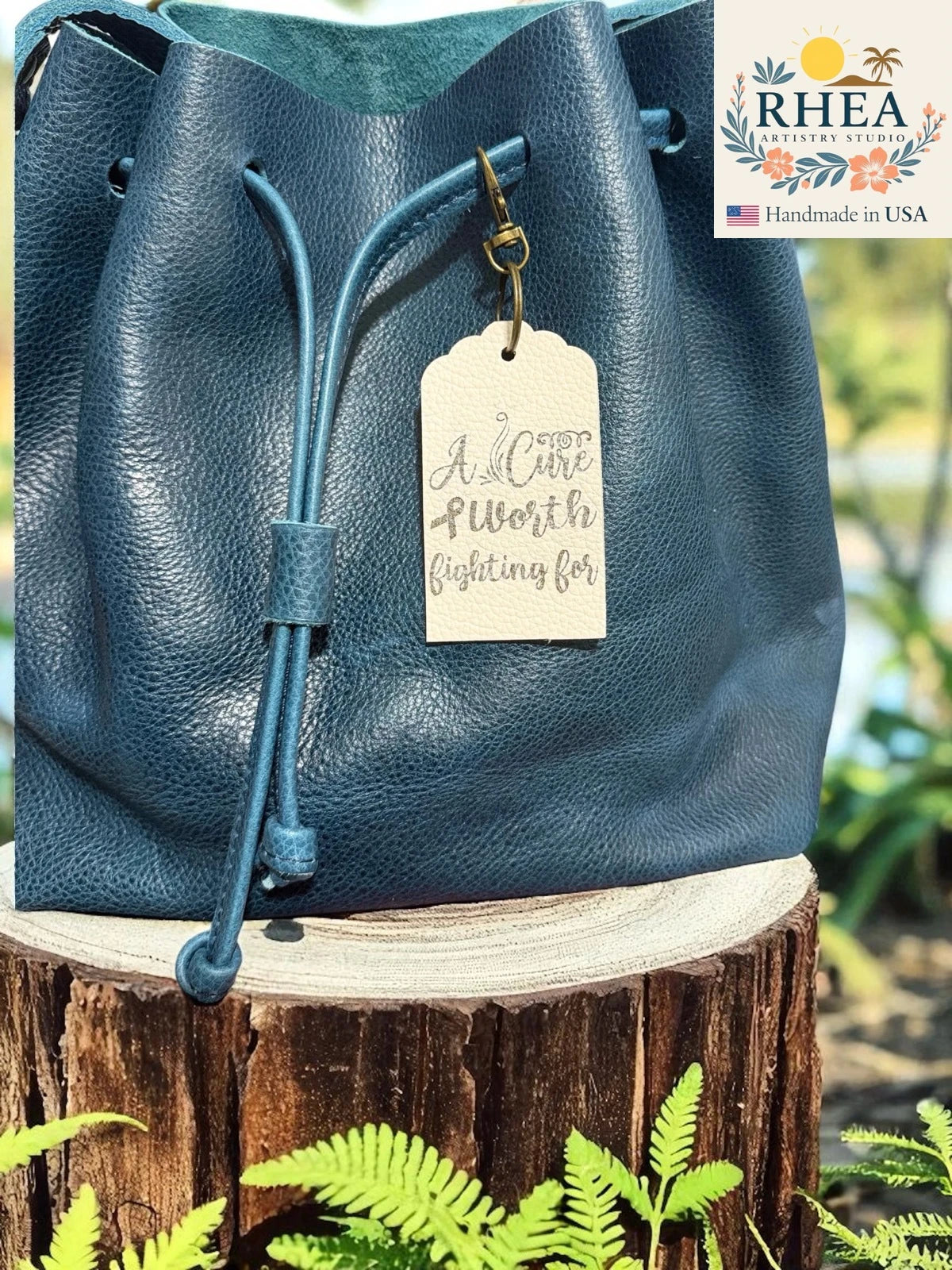 Blue leather bag with a leather tag on a wooden stump with plants in the background, featuring the RHEA brand.