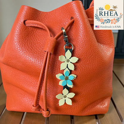 Orange leather bag with a floral keychain charm on a wooden surface, featuring the RHEA brand.