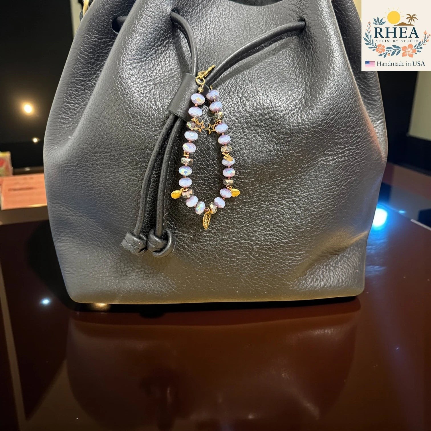 Gray leather drawstring bag with a purse charm on a reflective surface, RHEA brand visible.