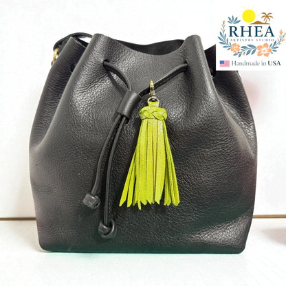 Black leather bucket bag with a green tassel on a white background, featuring the RHEA brand logo.