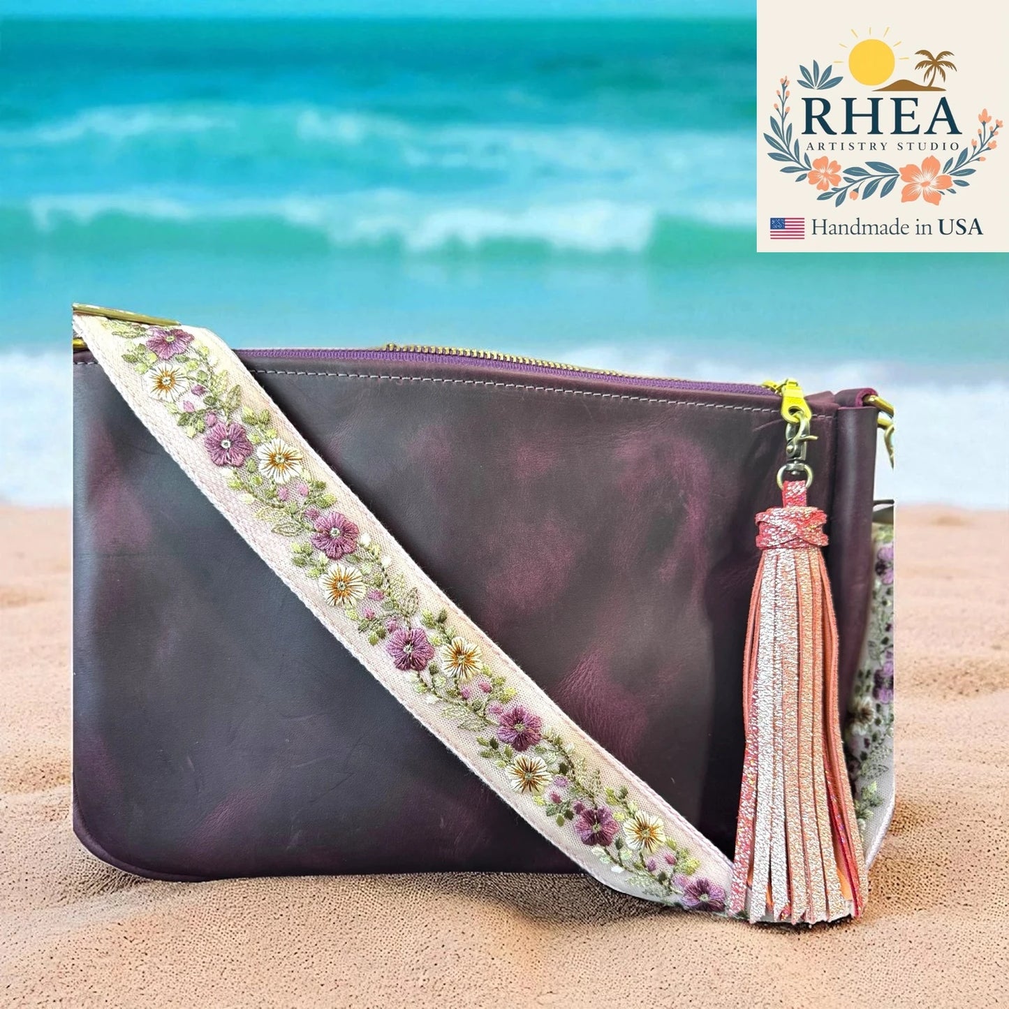 Handbag with floral leather bag strap and tassel on a beach, featuring RHEA brand.