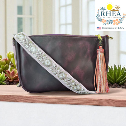 Purple handbag with luxury artisan strap and tassel on a wooden surface, RHEA brand visible.