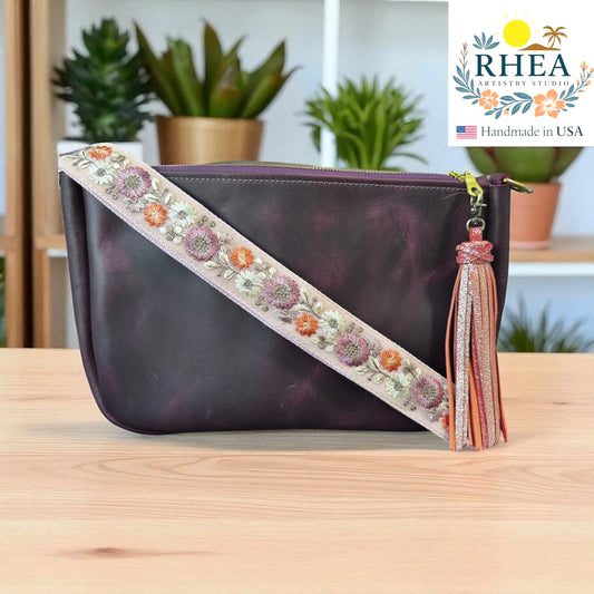 Purple handbag with floral strap and tassel on a wooden surface, RHEA brand visible.