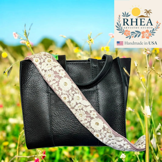 Black handbag with a floral purse strap in a field of flowers, featuring RHEA Artistry Studio branding.