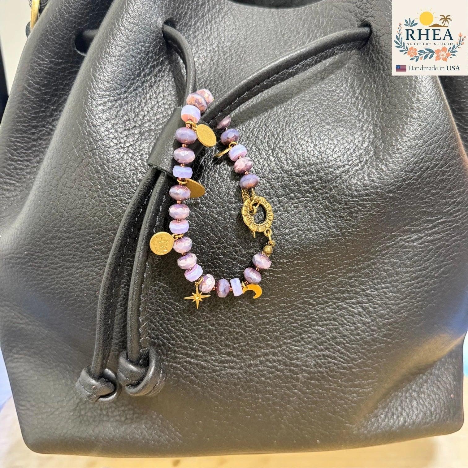 Bag charm with pink beads on a black leather bag, featuring the RHEA brand.
