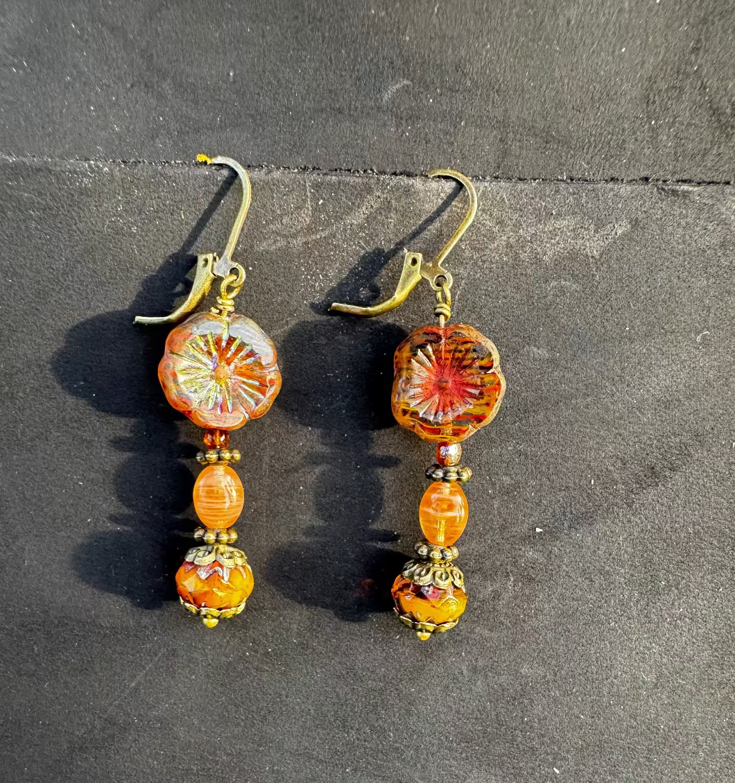 A pair of mustard yellow glass hibiscus flower drop earrings with brass accents placed on a dark surface.