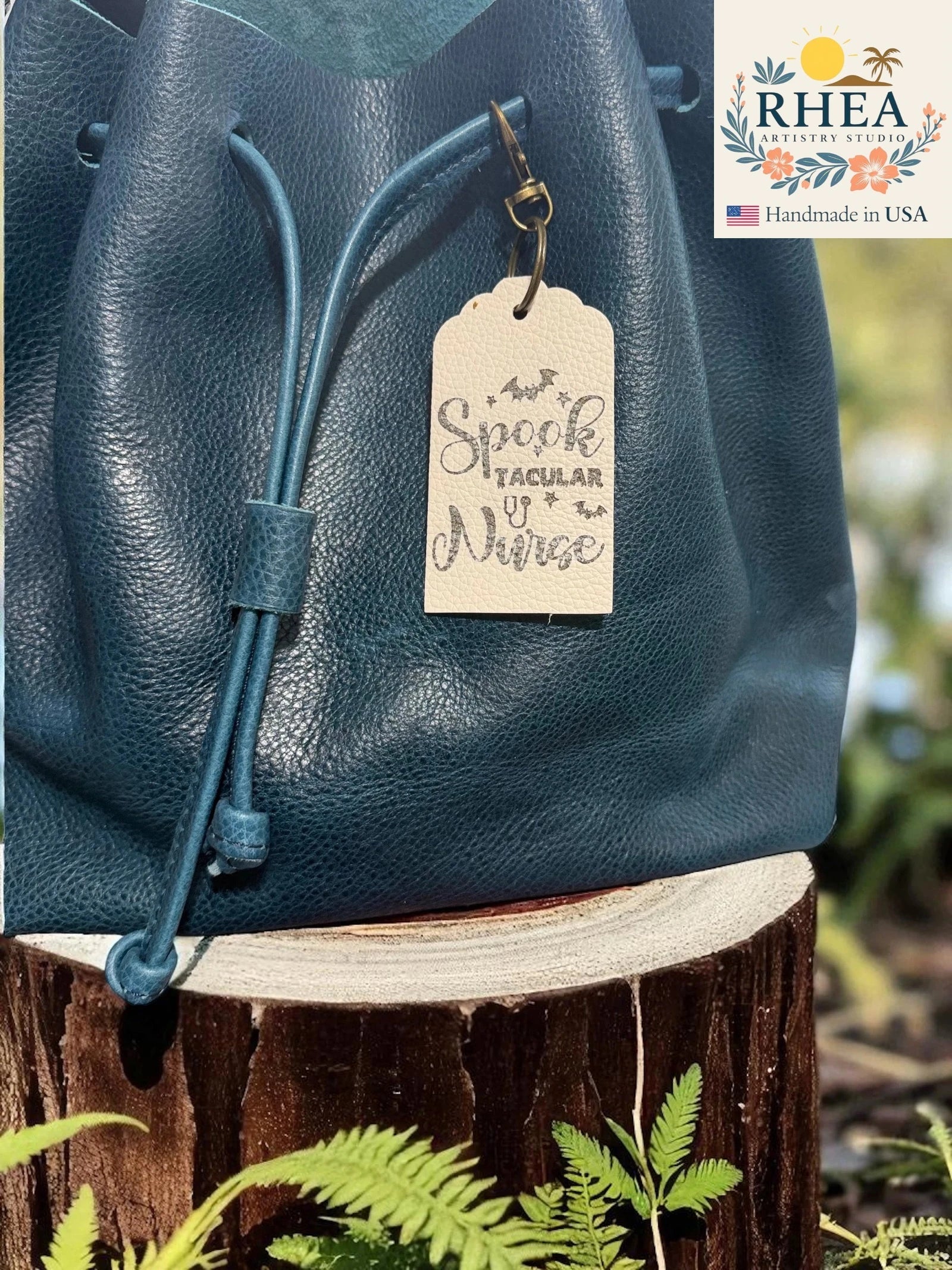 Blue leather bag with a decorative leather tag on a wooden stump with plants in the background, featuring the RHEA brand.