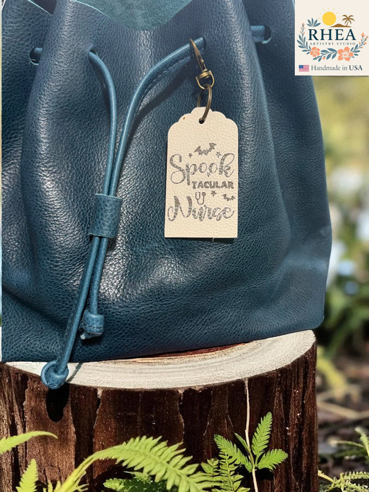 Blue leather bag with a leather engraved tag on a wooden stump with plants in the background, featuring the RHEEA brand.